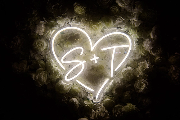 Wedding neon sign with a custom wedding neon sign initials and heart glowing over a white rose and hydrangea floral wall backdrop