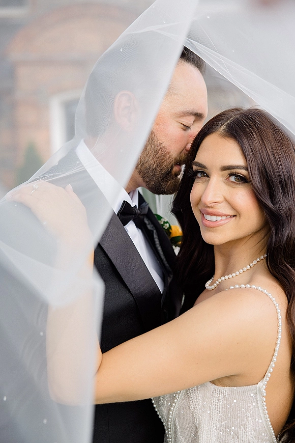 Couple portrait under a wedding veil, groom kissing bride’s forehead as she smiles at the camera by a brick doorway outdoors