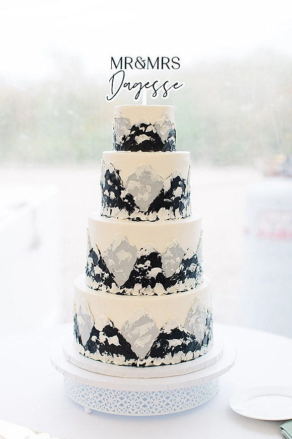 Wedding cake with mountain wedding cake design, four tiers of white frosting with black and gray icing on a stand by blurred windows