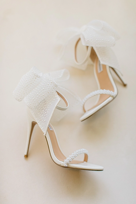 Wedding shoes in a bridal high heel sandals flat lay, white stilettos with pearl strap detail and tulle bows on a neutral floor