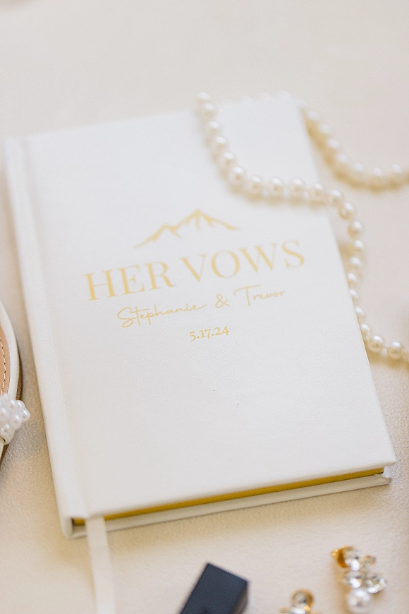 Wedding vows book with a ribbon bookmark beside a pearl necklace on a linen surface, styled as classic bridal detail flat lay