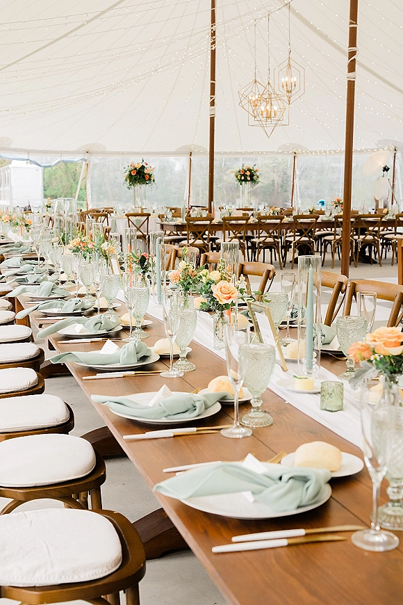 Reception tablescape on a long farm table wedding with sage green napkins, orange rose centerpieces, candles, and goblets under a clear tent