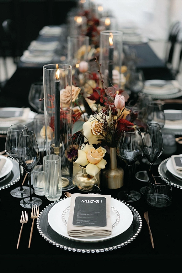 Reception tablescape with black wedding tablescape details—rose centerpiece, taper candles in glass hurricanes, and elegant place settings on a long banquet table