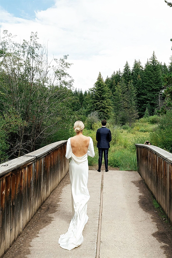 First look moment as bride in a low-back gown with train walks to groom in a suit on a wooden bridge amid forest greenery