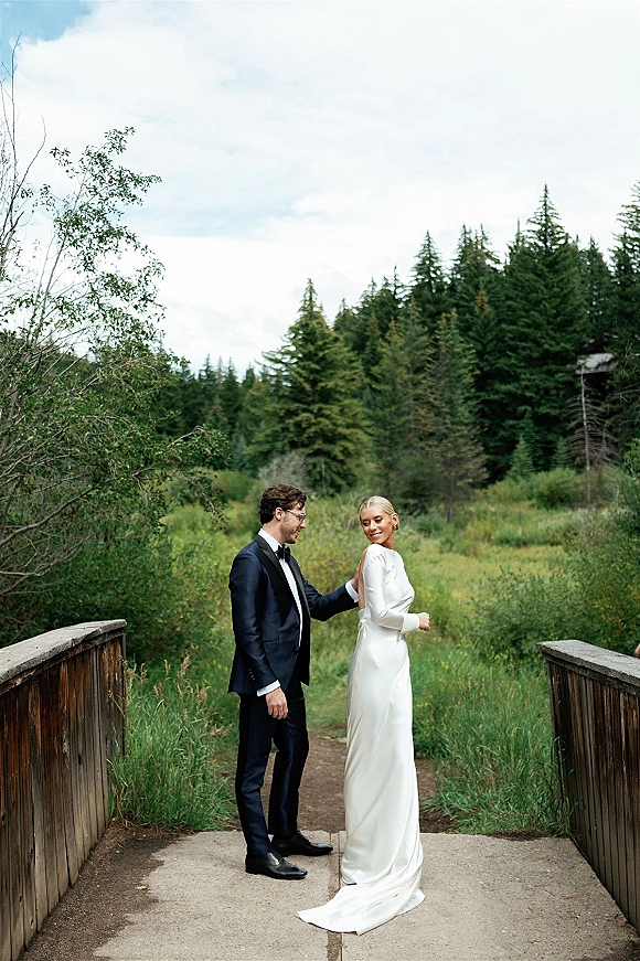 Couple portrait of bride in long-sleeve wedding dress and groom in tuxedo on a wooden bridge, with meadow and evergreen trees behind them