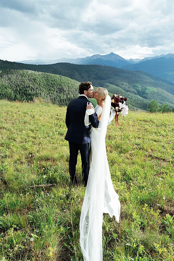 Wedding kiss portrait of bride and groom kissing, veil flowing as she holds a burgundy bouquet in a mountain meadow under cloudy skies