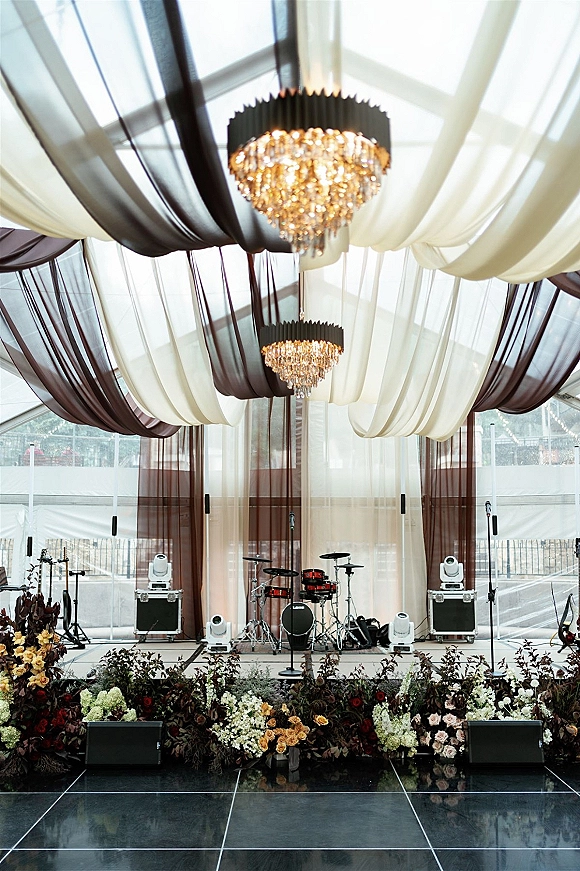 Reception stage decor with wedding band stage setup, drum kit and microphones under crystal chandeliers in a clear glass tent