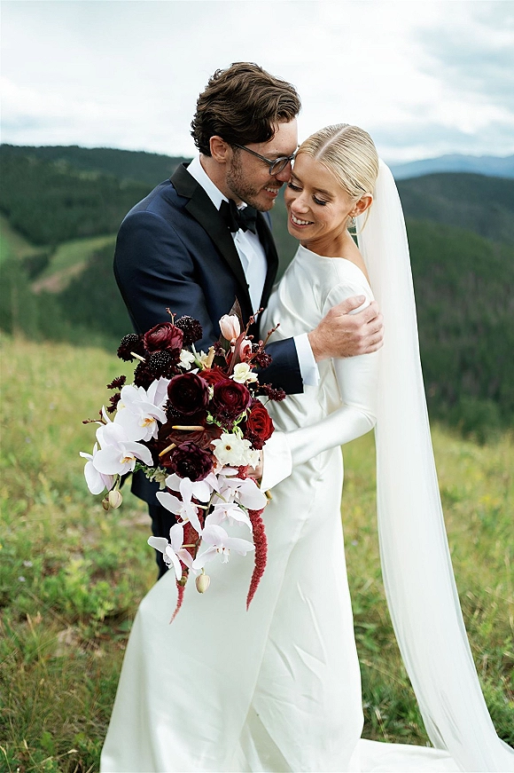 Couple portrait of bride and groom touching foreheads in an alpine meadow, bride with cathedral veil and cascading orchid bouquet, mountains behind
