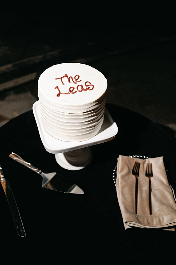 Wedding cake on a square stand with piped icing lettering, with cake knife, server, forks and folded napkins on a black tablecloth