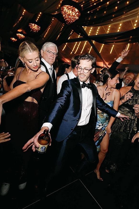 Wedding dance floor packed with guests dancing under string lights and chandeliers in a reception tent, tuxedos and sequin dresses holding drinks