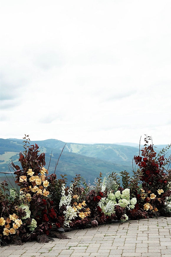Wedding floral installation with roses, white blooms and greenery with burgundy foliage lining brick pavers against a cloudy mountain view