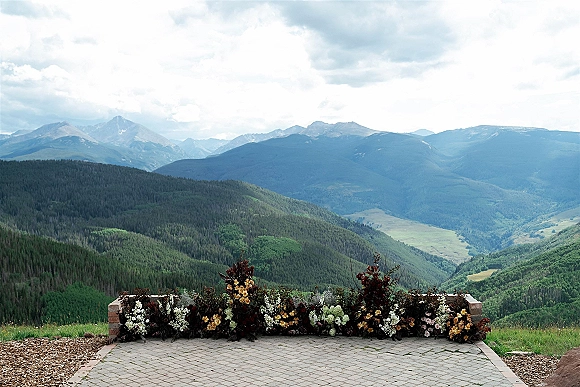 Wedding altar flowers in a grounded altar florals installation with roses and greenery on stone pavers, set before misty mountain hills and valley