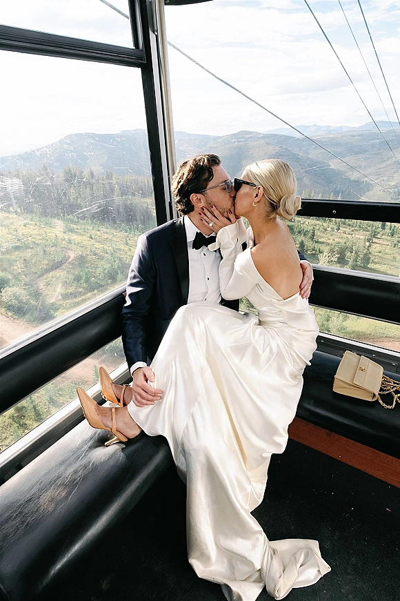 Wedding kiss portrait in a gondola wedding photo, bride in sunglasses and satin gown kissing groom in tux, mountains beyond windows