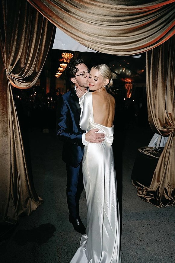 Wedding couple portrait with groom kissing bride’s cheek as they embrace in a dim reception with chandeliers, bistro lights, and draped curtains