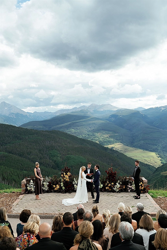 Wedding ceremony at an outdoor mountain ceremony overlook as the couple exchanges vows under a floral arch with guests and cloudy peaks behind
