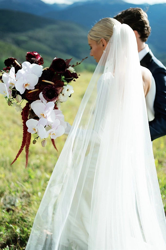 Couple portrait of bride and groom with a long veil and cascading orchid bouquet, standing in a grassy mountain meadow under blue sky