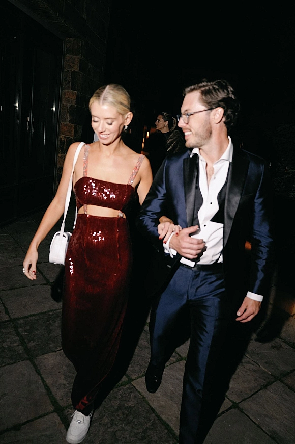 Wedding guest style couple walking arm in arm at night, woman in burgundy sequin gown with cutout and white shoulder bag, man in navy tuxedo
