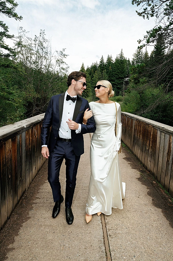 Couple portrait of bride and groom walking arm in arm on a wooden bridge, wearing sunglasses, her gown train trailing behind