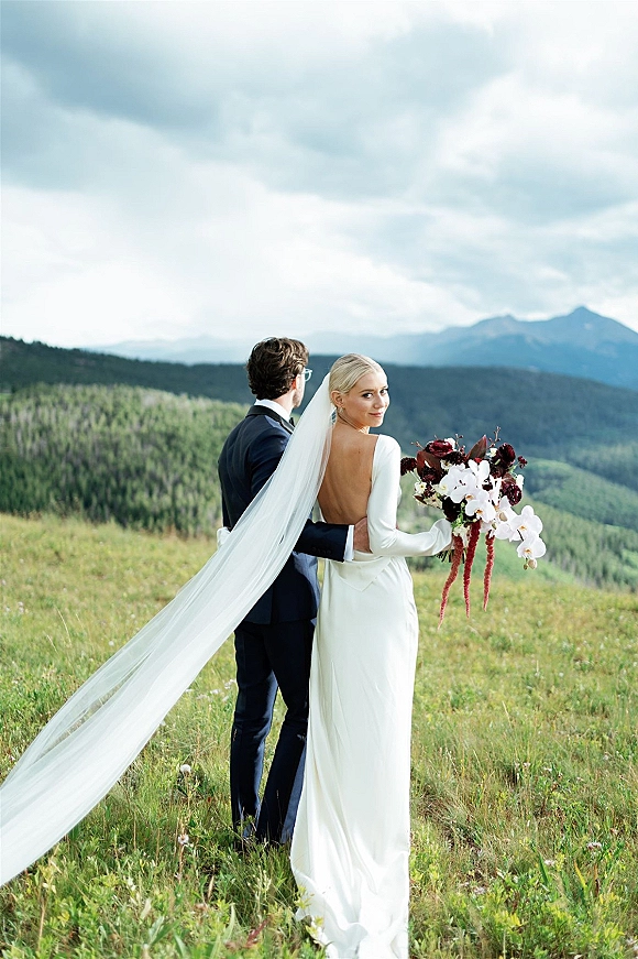 Couple portrait in a mountain wedding portrait, bride in long veil and gown holding bouquet as groom in tux embraces her in meadow grass