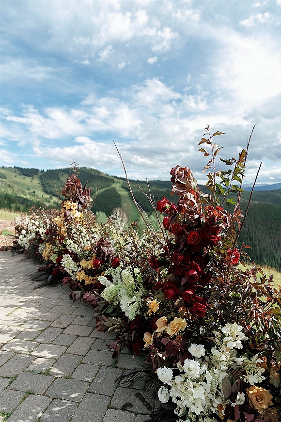 Ceremony aisle flowers line a stone paver walkway, with red and cream roses, greenery, and burgundy foliage against mountain hills