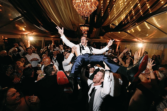 Wedding reception dance as guests cheer during a hora chair lift, arms raised under a chandelier in a draped-ceiling reception tent at night