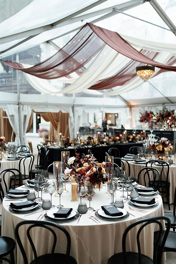 Reception tablescape with round wedding reception tables in beige linens, black chairs, floral centerpieces and taper candles under a clear top tent