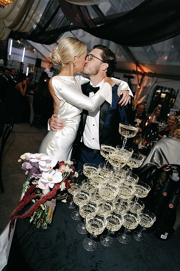 Wedding kiss as bride in a satin gown and groom in tuxedo and glasses kiss beside a champagne coupe tower under draped tent ceiling lights