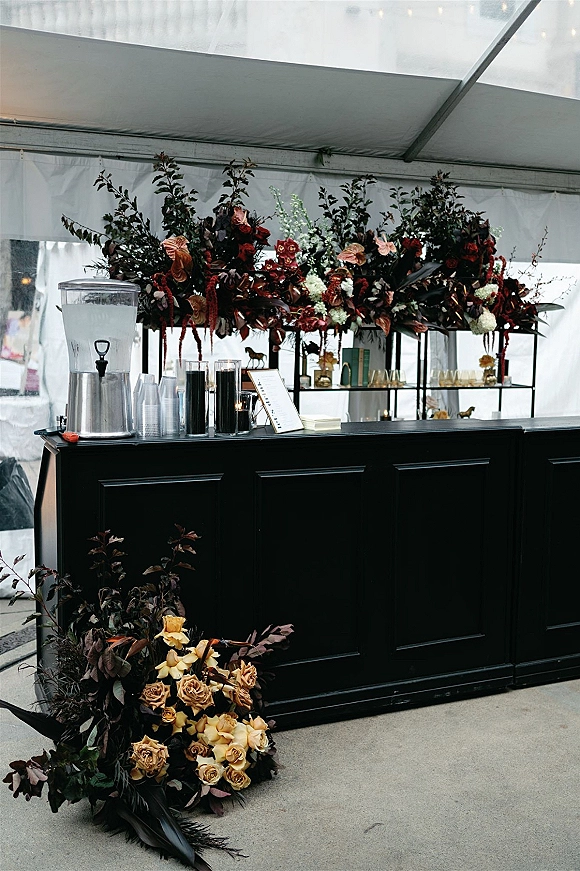 Wedding bar setup featuring a black mobile wedding bar with tall floral centerpiece, candles, bud vases, and menu sign in a clear tent