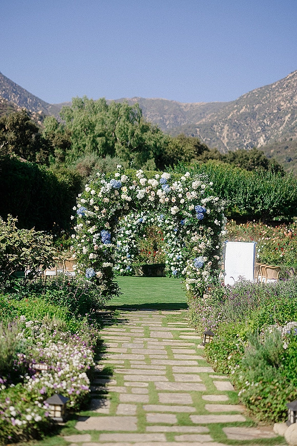 Ceremony aisle design featuring an outdoor wedding aisle of stone pavers lined with lanterns, hydrangea rose arch, and mountain lawn backdrop