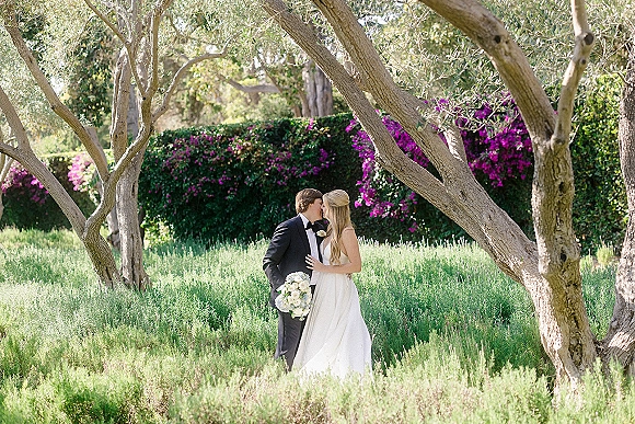 Wedding kiss as the bride holds a white rose bouquet, wearing a strapless gown beside the groom in a black tux, framed by garden greenery