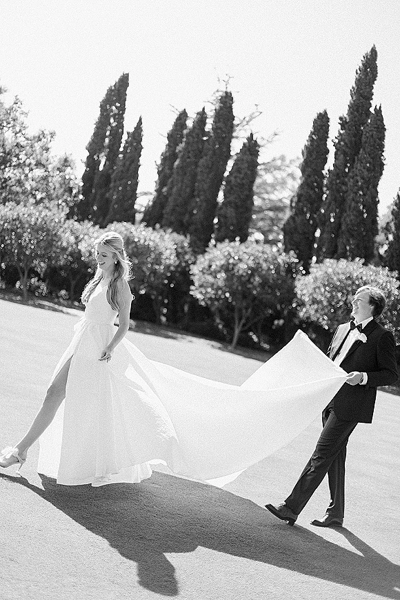 Couple portrait of bride and groom walking as he holds her wedding dress train, captured on a sunlit road lined with hedges and tall trees