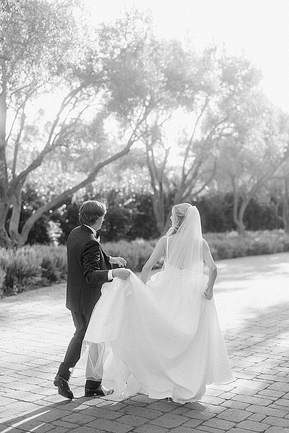Wedding couple walking down a tree-lined path, bride and groom walking away in tuxedo and long veil, sunlight on paving stones