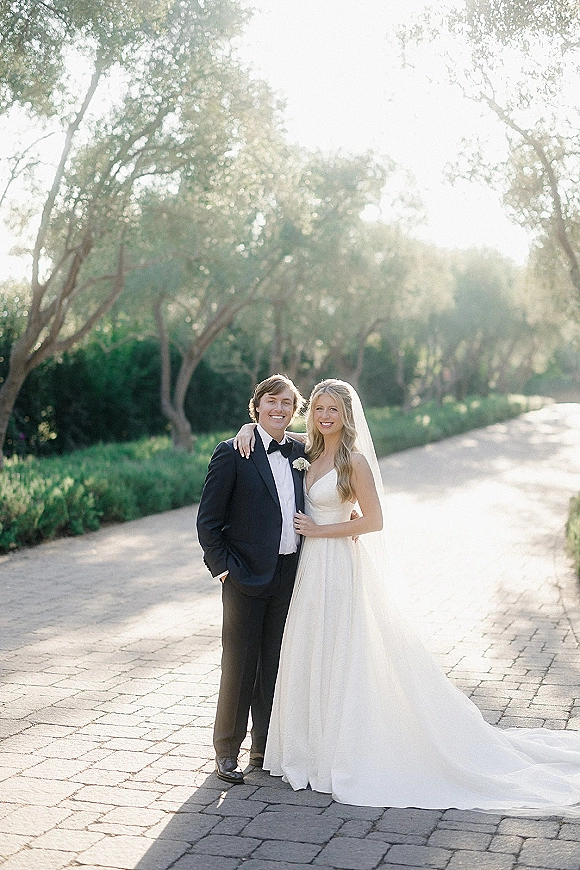 Couple portrait of bride and groom smiling on a sunlit tree-lined path, bride in veil with arm around groom in tuxedo