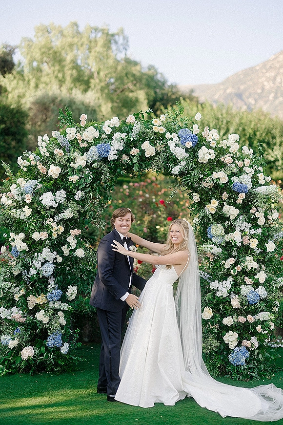 Couple portrait of bride and groom smiling under a circle floral arch with white roses and blue hydrangeas in a mountain garden setting