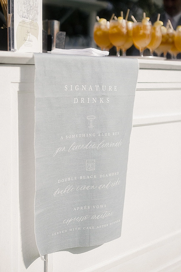 Signature drinks sign with wedding bar sign calligraphy on linen, displayed on an acrylic stand beside cocktail glasses on a white bar counter