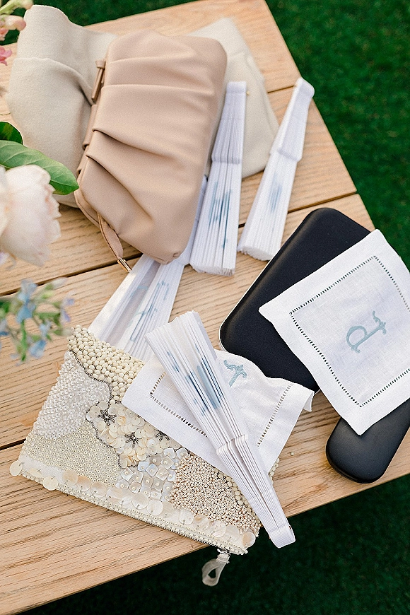 Bridal accessories flat lay of wedding day essentials including beaded, blush and black clutches, folding fans, monogram napkin and flowers on a wooden table outdoors