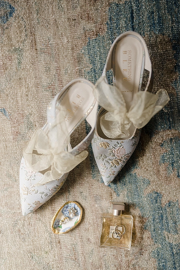 Bridal shoes in a wedding shoe flatlay, white embroidered slingback heels with tulle bows beside a ring and perfume on a patterned rug