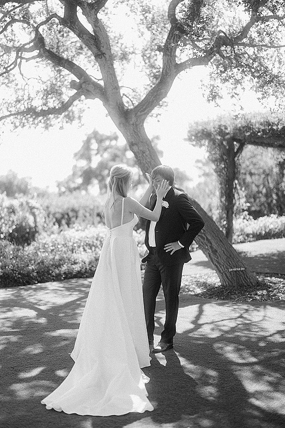 Couple portrait in a black and white wedding portrait under a large garden tree, bride in veil touching groom’s face in sunlight