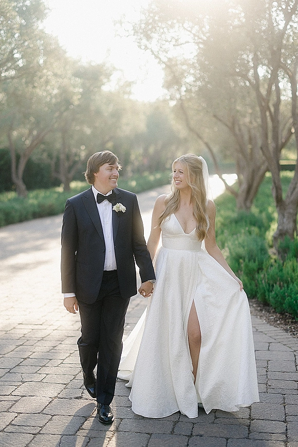 Couple portrait of bride and groom walking hand in hand on a sunlit tree-lined path, her veil flowing beside his black tuxedo