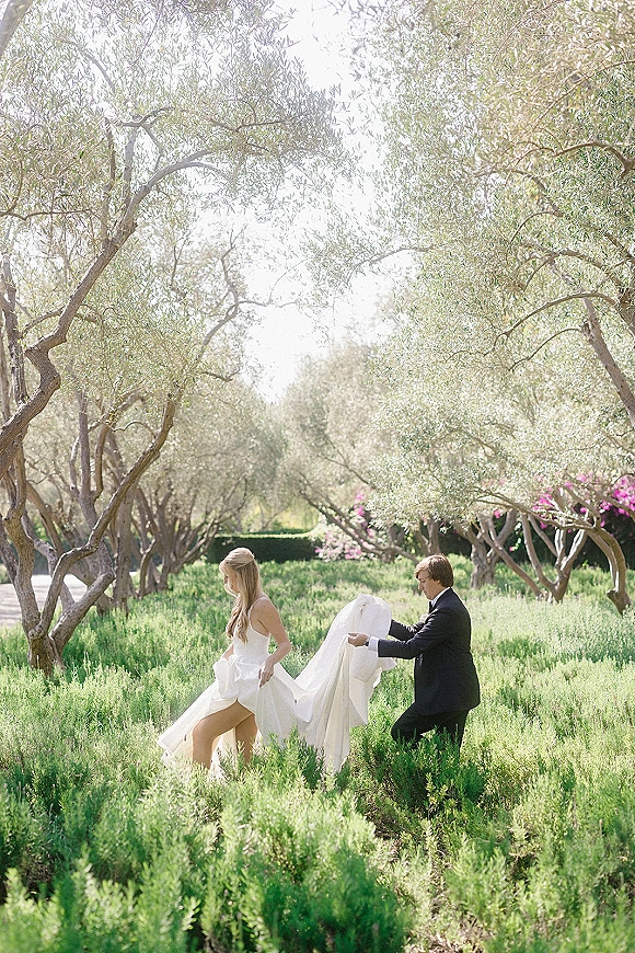 Couple portrait of bride and groom walking as groom holds the wedding dress train through an olive grove meadow with flowering bushes