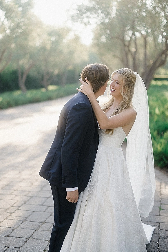 Wedding couple portrait of bride and groom embrace as she smiles, long veil flowing, on a sunlit tree-lined path with stone pavers