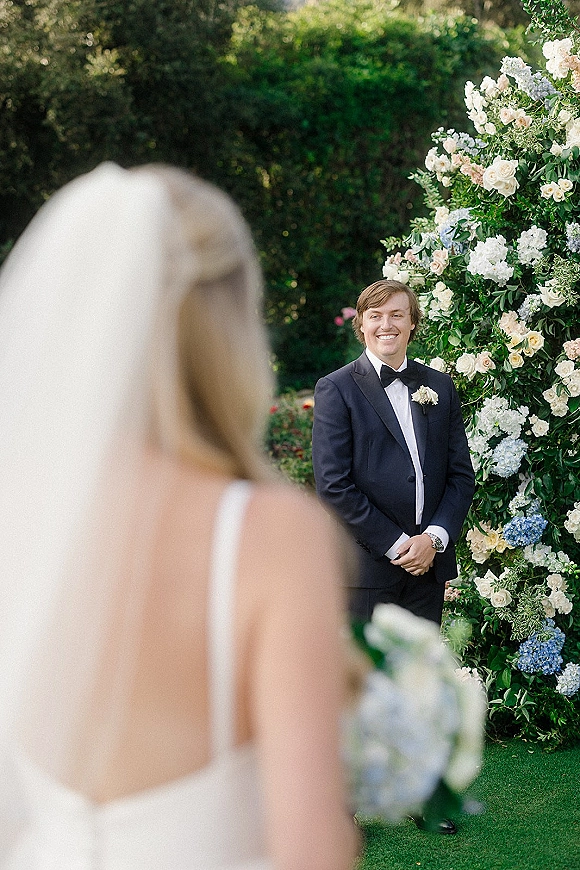 Wedding first look as bride in veil walks up behind groom in tuxedo holding a bouquet, set by garden hedges and a floral wall