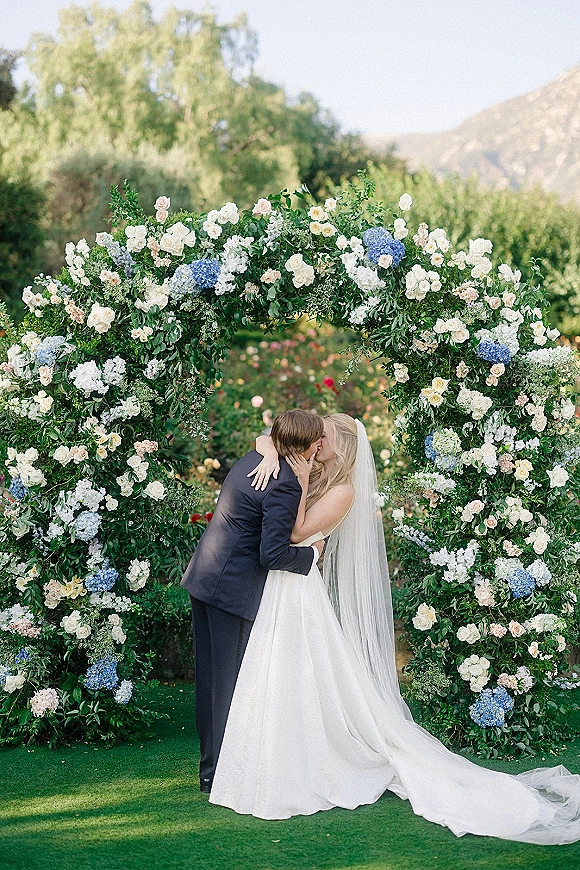 Wedding kiss portrait of bride and groom kissing under arch with a long veil, framed by roses and hydrangeas in a garden with mountains