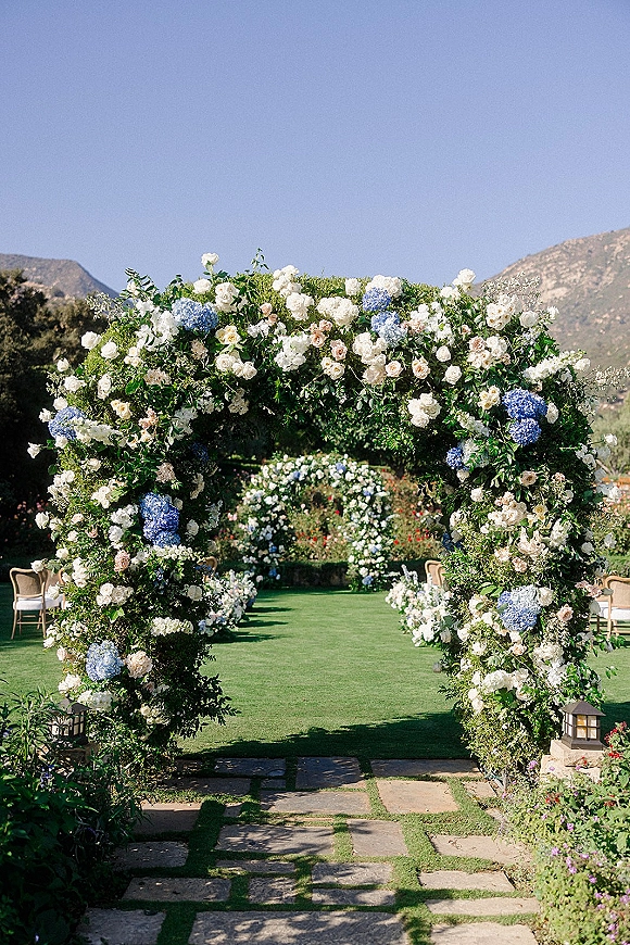 Wedding ceremony arch adorned with roses and hydrangeas over a stone walkway aisle, with garden lanterns and mountains beyond