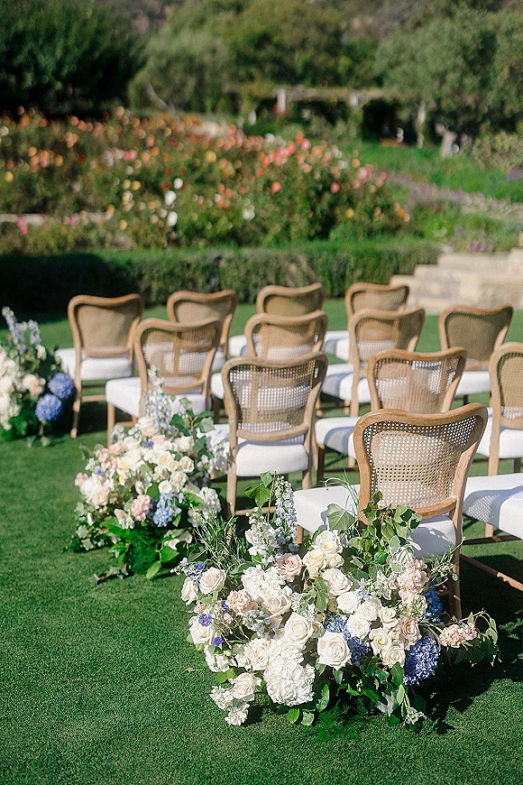 Ceremony setup with outdoor ceremony seating on a garden lawn, wood cane chairs and lush roses, hydrangea, and greenery lining the aisle