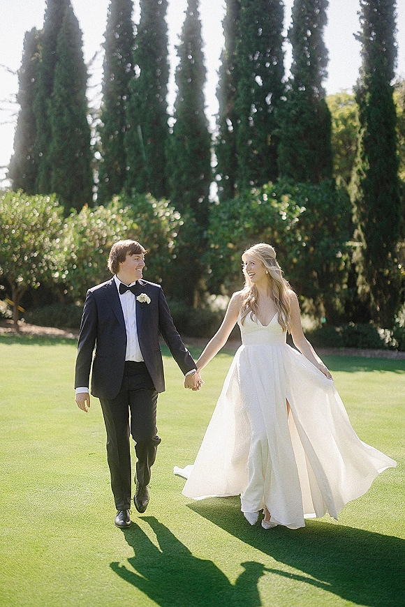 Couple portrait of bride and groom walking hand in hand, bride twirling her veil, on a sunlit lawn with tall trees and hedges
