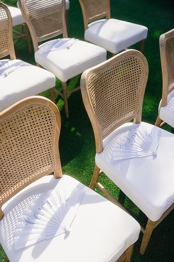 Ceremony seating with outdoor ceremony chairs featuring cane backs, white cushions, and paper hand fans on a sunlit green lawn