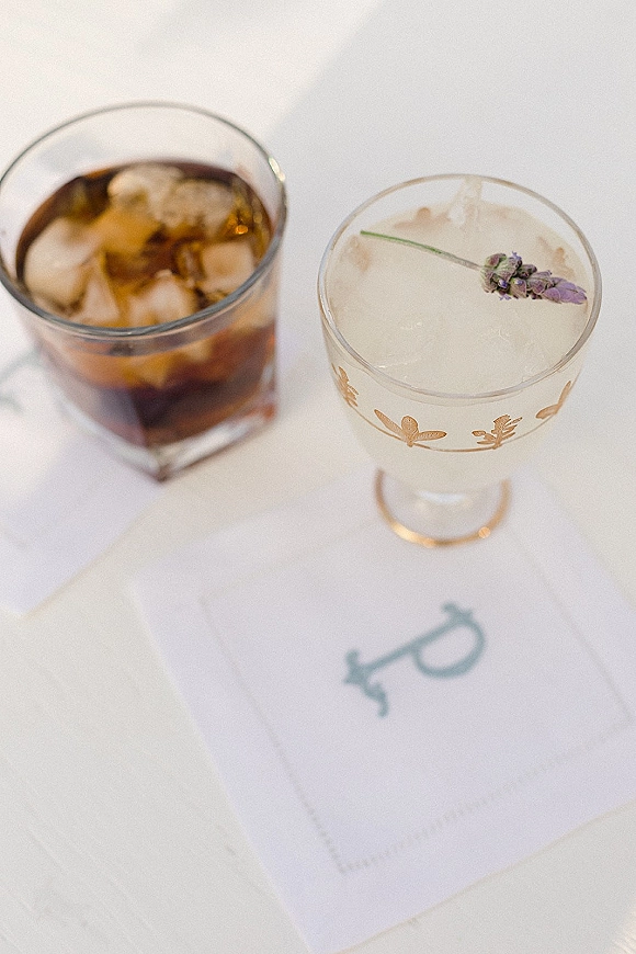 Wedding cocktails with a lavender sprig and ice in gold-rimmed glasses beside a printed menu card on a white tablecloth flatlay