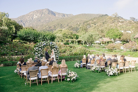 Outdoor wedding ceremony with bride and groom beneath a circular floral arch, wooden chairs on a manicured lawn with mountains behind