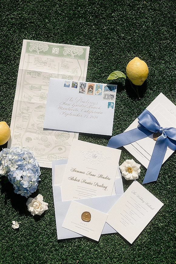 Wedding invitation suite with letterpress wedding invitations, script envelope, blue ribbon and wax seal, styled with lemon and hydrangea on grass